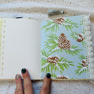 K&ouml;nnte beinhalten: Ein offenes Journal mit einer leeren wei&szlig;en Seite links und einer blauen Seite mit einem Tannenzapfenmuster rechts. Das Journal hat ein gelbes Band und einen wei&szlig;en Spitzenbesatz.