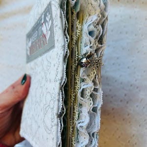 K&ouml;nnte beinhalten: Ein wei&szlig;es Stoffjournal mit einem silbernen und roten Etikett "Keep it Merry". Das Journal ist mit Spitze und einem silbernen Schneeflocken-Anh&auml;nger verziert.