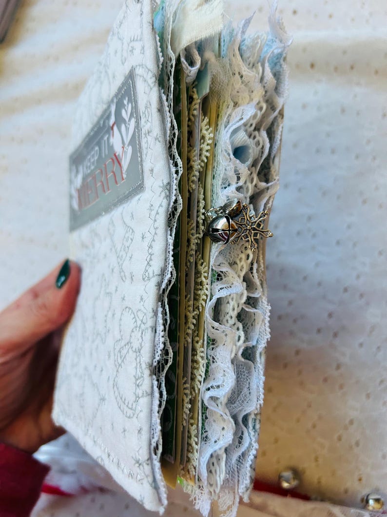 K&ouml;nnte beinhalten: Ein wei&szlig;es Stoffjournal mit einem silbernen und roten Etikett "Keep it Merry". Das Journal ist mit Spitze und einem silbernen Schneeflocken-Anh&auml;nger verziert.