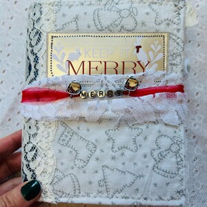 K&ouml;nnte beinhalten: Ein wei&szlig;es Stoffjournal mit einer Spitzenborte und einem goldenen Anh&auml;nger mit der Aufschrift "Keep it Merry". Das Journal ist mit rotem Band und Gl&ouml;ckchen verziert, auf denen das Wort "Merry" in Silber geschrieben steht.