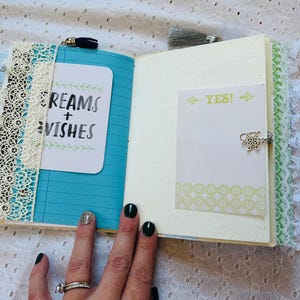 K&ouml;nnte beinhalten: Ein offenes Tagebuch mit einem blauen Einband und wei&szlig;er Spitzenverzierung. Die linke Seite hat eine wei&szlig;e Karte mit dem Text "Dreams + Wishes" in schwarzer Tinte geschrieben. Die rechte Seite hat eine wei&szlig;e Karte mit dem Text "YES!" in schwarzer Tinte geschrieben.