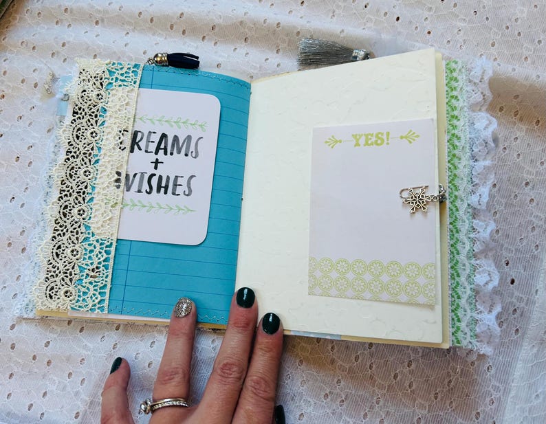 K&ouml;nnte beinhalten: Ein offenes Tagebuch mit einem blauen Einband und wei&szlig;er Spitzenverzierung. Die linke Seite hat eine wei&szlig;e Karte mit dem Text "Dreams + Wishes" in schwarzer Tinte geschrieben. Die rechte Seite hat eine wei&szlig;e Karte mit dem Text "YES!" in schwarzer Tinte geschrieben.