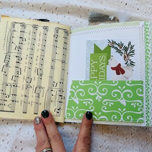 K&ouml;nnte beinhalten: Ein handgemachtes Journal mit einer wei&szlig;en Spitzenborte und einem gr&uuml;nen, gemusterten Papierumschlag. Das Journal ist ge&ouml;ffnet und zeigt eine Seite mit Noten und ein gr&uuml;nes, gemustertes Papier mit dem Text "Frohe Weihnachten".