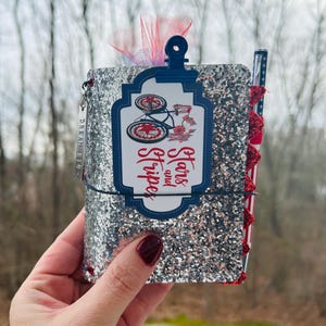 May include: A glittery silver notebook with a decorative tag featuring a bicycle and the words "Stars and Stripes." The notebook has a red and white ribbon and a pen with a patriotic design. A small charm reads "Dream Big."