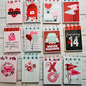 May include: A collection of Valentine's Day themed spiral-bound notepads. Each notepad features a unique design with hearts, flags, floral arrangements, and phrases like "Forever & Ever" and "Love You." The color palette includes red, pink, and white.