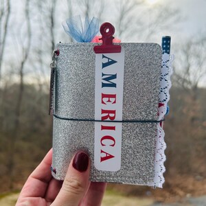 May include: A silver glitter journal with a red and blue "AMERICA" clip. The journal is secured with a black elastic band and has a white lace trim. A blue tulle bow and a small charm are attached.