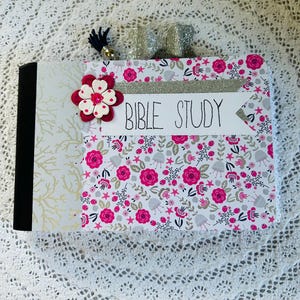 May include: A white notebook with a pink floral pattern and a black spine. The front cover has a silver glitter ribbon, a pink flower with a white center, and a white label with the text "Bible Study".
