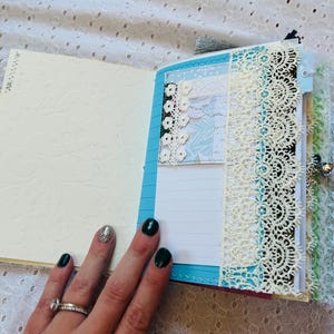 K&ouml;nnte beinhalten: Ein blaues und wei&szlig;es Journal mit Spitzenbesatz und einem dekorativen Papier-Verzierst&uuml;ck. Das Journal ist auf einer leeren Seite mit liniertem Papier ge&ouml;ffnet.