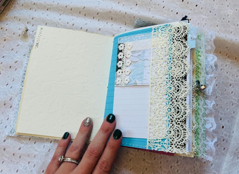 K&ouml;nnte beinhalten: Ein blaues und wei&szlig;es Journal mit Spitzenbesatz und einem dekorativen Papier-Verzierst&uuml;ck. Das Journal ist auf einer leeren Seite mit liniertem Papier ge&ouml;ffnet.