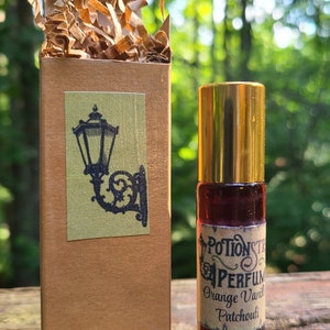 K&ouml;nnte beinhalten: Eine kleine braune Schachtel mit einem goldenen Aufkleber, der eine schwarze Silhouette einer Stra&szlig;enlaterne zeigt. Neben der Schachtel befindet sich eine kleine Glasflasche mit einem goldenen Verschluss und einem Etikett mit der Aufschrift "Potionstr Perfumes Orange Vanilla Patchouli".