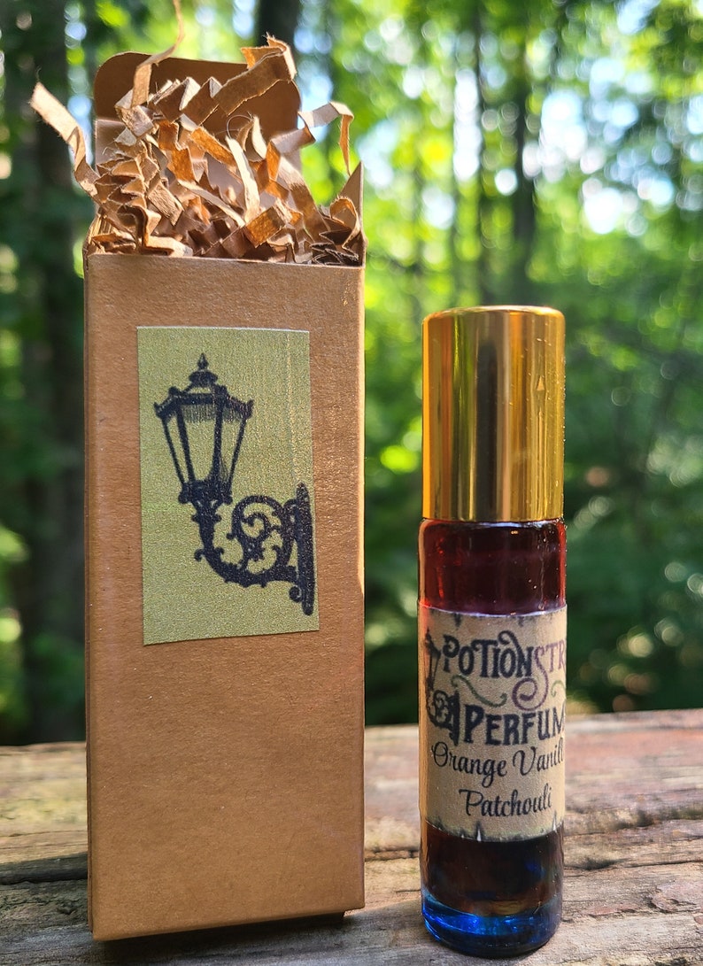 K&ouml;nnte beinhalten: Eine kleine braune Schachtel mit einem goldenen Aufkleber, der eine schwarze Silhouette einer Stra&szlig;enlaterne zeigt. Neben der Schachtel befindet sich eine kleine Glasflasche mit einem goldenen Verschluss und einem Etikett mit der Aufschrift "Potionstr Perfumes Orange Vanilla Patchouli".
