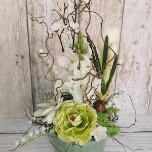 May include: A floral arrangement in a light green ceramic bowl. The arrangement features white flowers, a large green peony-like bloom, and decorative twigs. The background is a light wood-grain pattern.