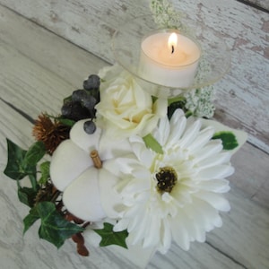 Puede incluir: Una decoración de mesa de temporada con una vela encendida en un soporte de vidrio, flores blancas, una calabaza blanca y follaje. El arreglo incluye una margarita blanca y hojas de hiedra.