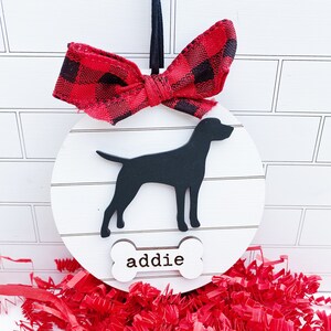 May include: A round, white wooden dog ornament with a black silhouette of a dog and the name "addie" on a bone-shaped tag. It is topped with a red and black buffalo plaid bow and a black ribbon for hanging. Red paper shreds are at the bottom.