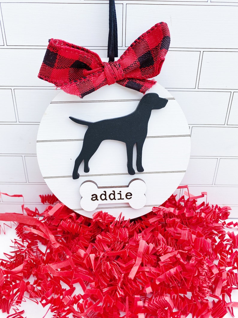 May include: A round, white wooden dog ornament with a black silhouette of a dog and the name "addie" on a bone-shaped tag. It is topped with a red and black buffalo plaid bow and a black ribbon for hanging. Red paper shreds are at the bottom.