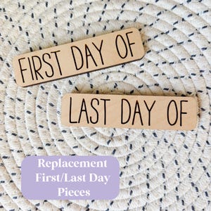 May include: Two rectangular wooden signs with the text "FIRST DAY OF" and "LAST DAY OF" in black. A light purple rectangle below reads "Replacement First/Last Day Pieces". The signs are on a white and beige woven surface.