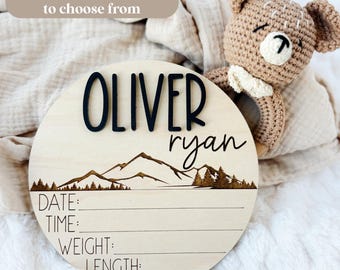 May include: A round wooden sign with the name "OLIVER" in black raised letters and "ryan" in script. The sign features a mountain and forest design, with lines for date, time, weight, and length. A crocheted bear toy is next to the sign.