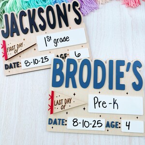 Puede incluir: Dos letreros escolares de madera, uno con "Jackson's" con "1st grade" y "Date: 8-10-25", el otro "Brodie's" con "Pre-K" y "Date: 8-10-25". Ambos tienen detalles de edad y bander&iacute;n. Una colorida guirnalda de borlas en el fondo.