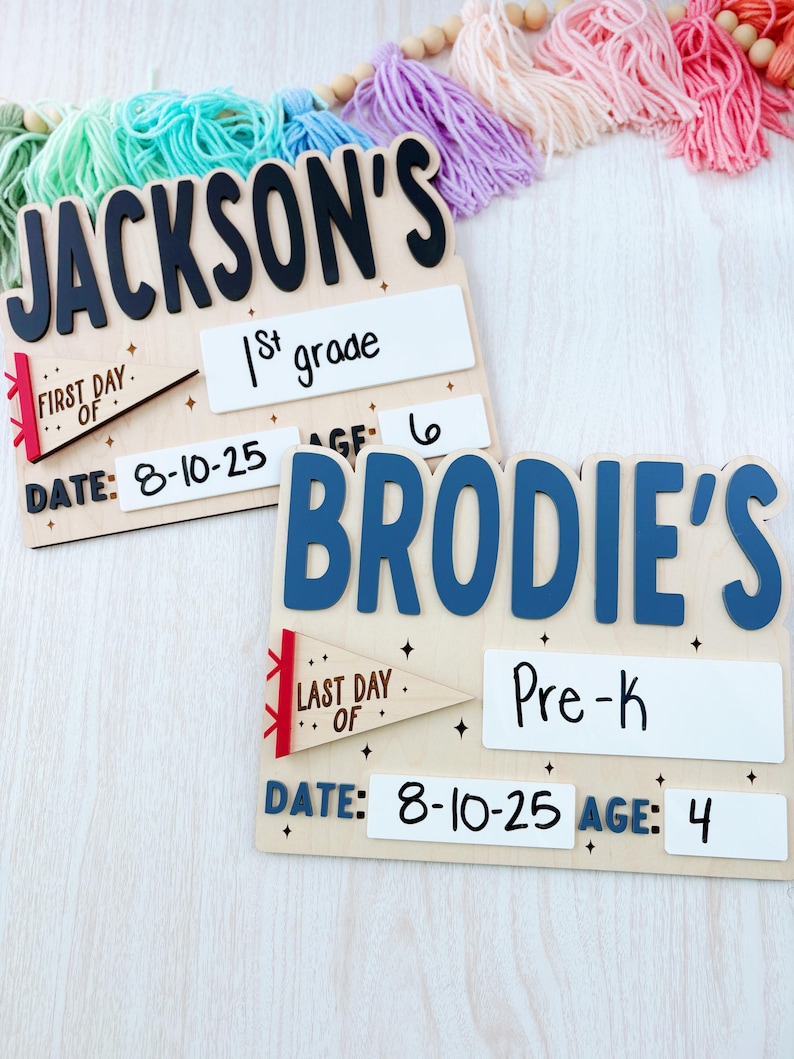 Puede incluir: Dos letreros escolares de madera, uno con "Jackson's" con "1st grade" y "Date: 8-10-25", el otro "Brodie's" con "Pre-K" y "Date: 8-10-25". Ambos tienen detalles de edad y bander&iacute;n. Una colorida guirnalda de borlas en el fondo.