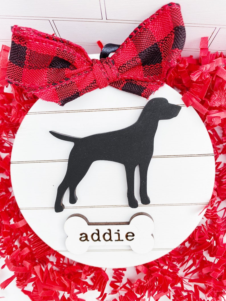 May include: A white wooden dog ornament with a black silhouette of a dog and the name "addie" on a bone-shaped tag. The ornament has a red and black plaid bow and is surrounded by red paper shreds.