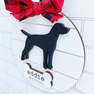 May include: A round, wooden dog ornament with a black silhouette of a dog and the name "addie" on a bone-shaped tag. The ornament is decorated with a red and black plaid bow and a black ribbon for hanging.