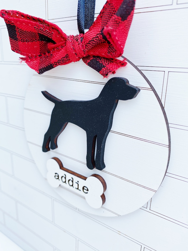 May include: A round, wooden dog ornament with a black silhouette of a dog and the name "addie" on a bone-shaped tag. The ornament is decorated with a red and black plaid bow and a black ribbon for hanging.