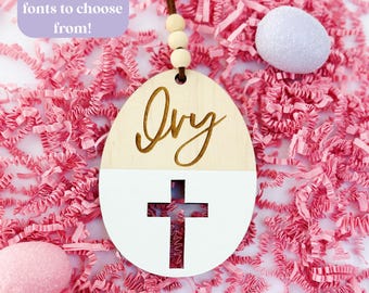 May include: An Easter egg-shaped wooden ornament with the name "Ivy" in a cursive font. The bottom half is white with a cut-out cross. The ornament hangs from a brown cord with wooden beads. Text reads "6 colors and 10 fonts to choose from!"