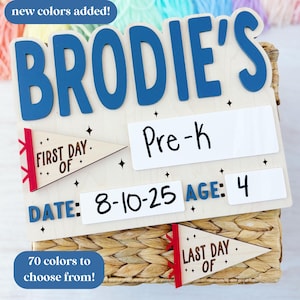 This photo features a pennant flag themed first and last day of school sign. The top of the sign is personalized with your child's name. font, and color of your choosing. There are interchangeable pennant flags that say First Day Of and Last Day Of with a customizable pennant flag ribbon color. The photo shows that there are 70 colors to choose from, with new colors recently added.