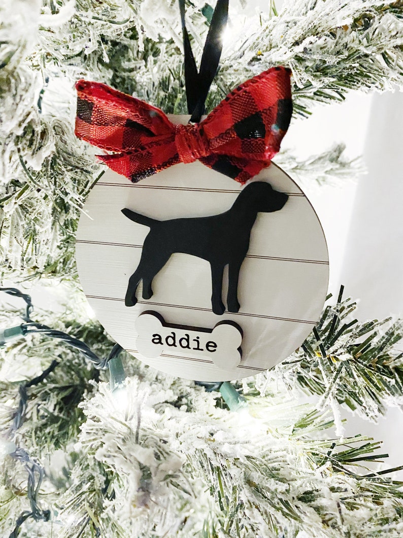 May include: A round, wooden Christmas ornament with a black dog silhouette and the name "addie" on a bone-shaped tag. The ornament has a red and black plaid bow and hangs from a black ribbon. The background is a snow-covered Christmas tree.