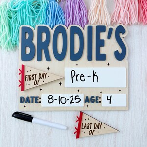 Puede incluir: Un letrero de madera con el nombre "BRODIE'S" en letras azules. Incluye secciones para "Pre-K", "Fecha: 8-10-25" y "Edad: 4". Banderines decorativos que dicen "First Day Of" y "Last Day Of". Se incluye un marcador negro. Borlas coloridas cuelgan arriba.