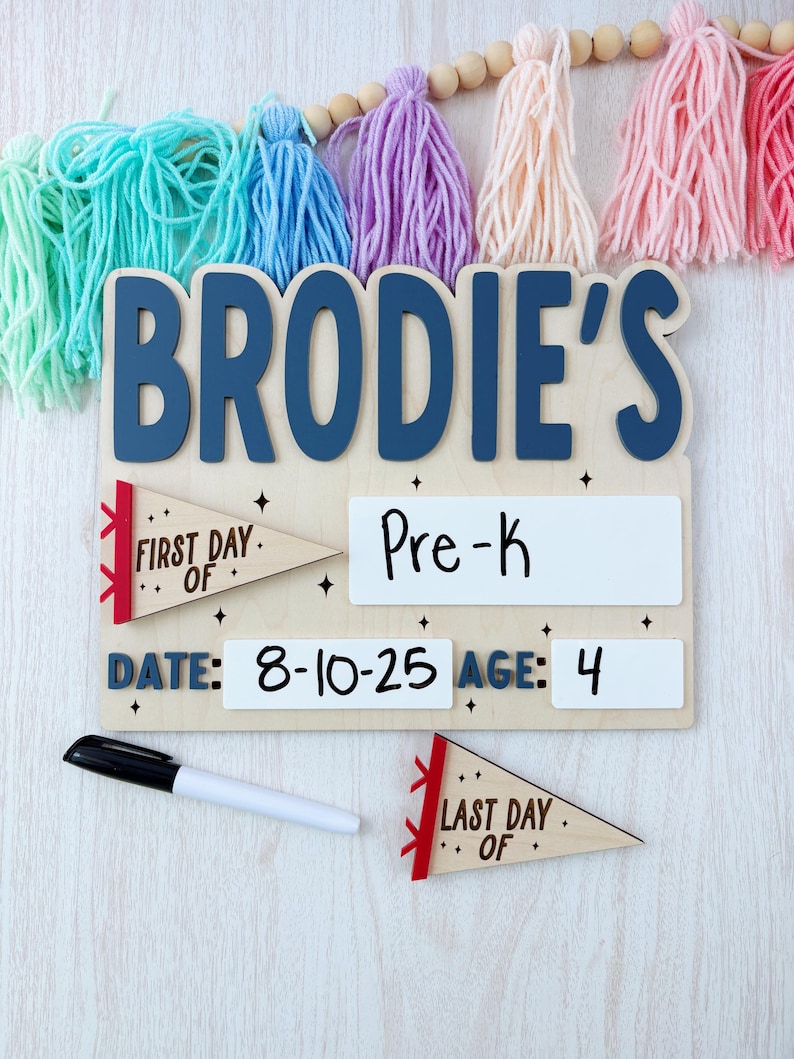 Puede incluir: Un letrero de madera con el nombre "BRODIE'S" en letras azules. Incluye secciones para "Pre-K", "Fecha: 8-10-25" y "Edad: 4". Banderines decorativos que dicen "First Day Of" y "Last Day Of". Se incluye un marcador negro. Borlas coloridas cuelgan arriba.