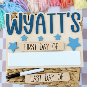 May include: A wooden sign with the name "WYATT'S" in blue letters, "FIRST DAY OF" and "LAST DAY OF" text, and blue star accents. Includes a white space for writing and a black marker. The sign is set on a woven basket.