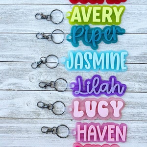 May include: Eight colorful keychains with different names on them, including Cooper, Avery, Piper, Jasmine, Lilah, Lucy, Haven, and Renee. Each keychain has a silver ring and a clear plastic backing.