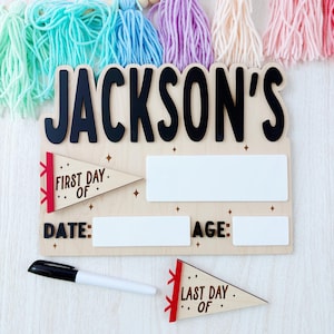 Puede incluir: Letrero de madera con el nombre "JACKSON'S" en letras negras. El letrero incluye espacios para "DATE" y "AGE", y detalles en forma de bander&iacute;n que dicen "FIRST DAY OF" y "LAST DAY OF". Se incluye un marcador negro.