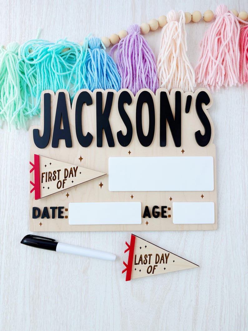 Puede incluir: Letrero de madera con el nombre "JACKSON'S" en letras negras. El letrero incluye espacios para "DATE" y "AGE", y detalles en forma de bander&iacute;n que dicen "FIRST DAY OF" y "LAST DAY OF". Se incluye un marcador negro.
