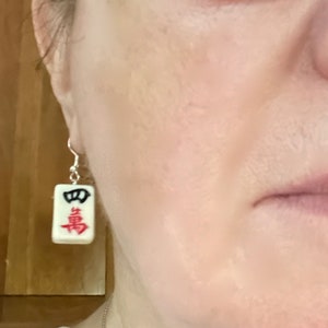 Puede incluir: Pendientes de cer&aacute;mica blanca con forma de ficha de mahjong con caracteres chinos negros y rojos. La ficha tiene un borde negro y los caracteres son rojos.
