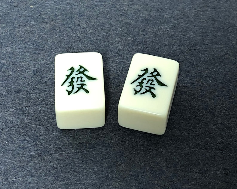 Puede incluir: Dos fichas blancas de mahjong con caracteres chinos negros en ellas. Los caracteres son "發", que significa "prosperidad" en ingl&eacute;s.