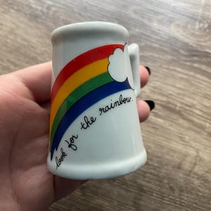 May include: White ceramic shot glass with a rainbow design in red, orange, yellow, green, and blue. The phrase "look for the rainbow" is written in black script. The glass has a small handle.
