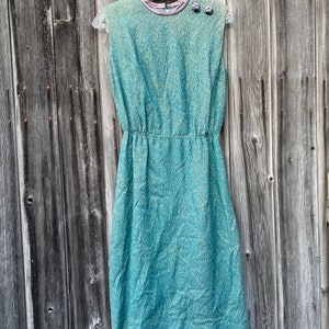 May include: A sleeveless teal blue dress with a textured pattern and a gathered waist. The dress has two small buttons on the shoulder.