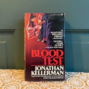 May include: A paperback book titled "Blood Test" by Jonathan Kellerman. The cover features a red and black color scheme with a close-up of a hand holding a bloody knife. The book is an Edgar Award-winning thriller.