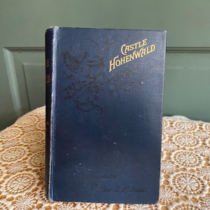 May include: A vintage book titled "Castle Hohenwald" with a dark blue cover. The title is in gold lettering, and there is an illustration of a face and floral design. The book is on a cream-colored lace tablecloth.