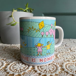 May include: White ceramic mug with a wraparound illustration of children flying kites in a park. The design includes a yellow kite, pink trees, and the text "LOVE IS HOME". The mug has a white handle.