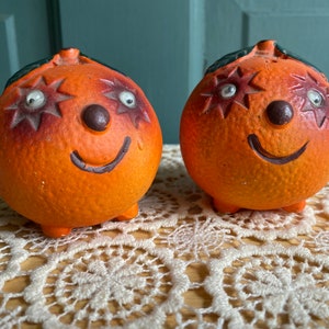 May include: Two orange ceramic salt and pepper shakers shaped like smiling oranges with black eyes and a black nose. The shakers have a star design on each side.