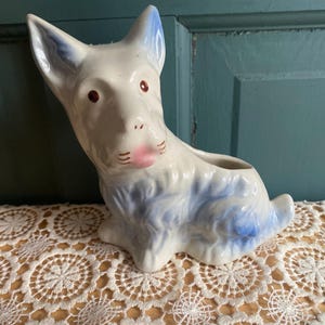 May include: A ceramic dog figurine, primarily white with blue accents on the ears and body. The dog has brown eyes and a pink mouth. The figurine is a planter, with an opening on its back.