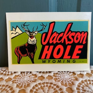 May include: A vintage postcard featuring a stylized illustration of a deer with large antlers in front of a green field and mountains. The text "Jackson Hole Wyoming" is in bold red letters.