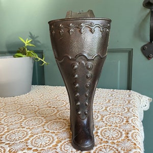 May include: A dark brown cast iron boot-shaped planter. The planter has a detailed design with decorative elements and button-like accents. It is placed on a cream-colored lace tablecloth, with a green door and a potted plant in the background.