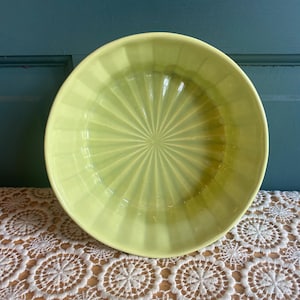 May include: A light green, round bowl with a ribbed, sunburst design radiating from the center. The bowl is made of ceramic or glass and sits on a white lace tablecloth. The background is a teal-colored wall.