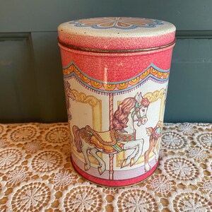 May include: A cylindrical metal storage tin featuring a carousel theme. The exterior displays a white background with illustrations of horses in shades of brown, gold, and red, topped with a pink lid decorated with a blue and orange floral pattern.
