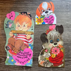 May include: Three vintage Valentine's Day cards featuring illustrations of a boy playing a harmonica, a dog with a heart, and a dog with a basket of flowers. The cards have pink, red, and blue accents and include the text "Perfect Harmony You & Me Valentine", "Valentine to say to be mine", and "With Love Happy Valentine's Day".