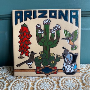 May include: A square ceramic tile with a cream background featuring the word ARIZONA in dark blue letters. The design includes a red chili pepper ristra, a green saguaro cactus with white flowers, a hummingbird, a roadrunner, and a pottery vessel.